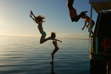 Young adult friends jumping from summer houseboat into sunset ocean