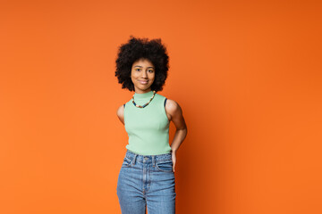 Smiling and stylish african american teenage girl in casual clothes with necklace and bold makeup posing and looking at camera isolated on orange, trendy teenage girl expressing individuality