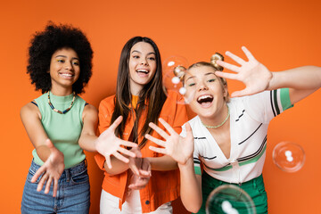 Excited and cheerful multiethnic teen girls with bold makeup looking at soap bubbles while posing...
