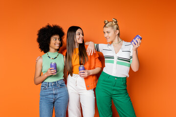 Positive blonde teenager in stylish outfit holding drink in tin can and looking at multiethnic girlfriends in casual outfits on orange background, trendy outfits and fashion-forward looks - Powered by Adobe