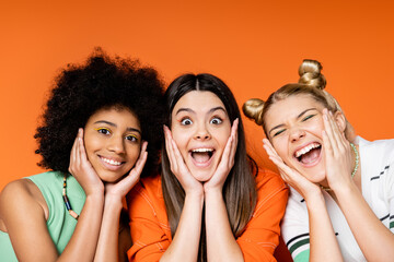 Excited and cheerful multiethnic teen girlfriends with bold makeup touching cheeks and looking at camera together on orange background, trendy outfits and fashion-forward looks, diverse races