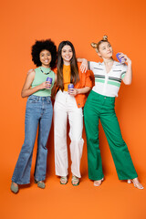 Fototapeta premium Full length of cheerful and stylish teenager hugging multiethnic girlfriends and holding canned drink while posing together on orange background, trendy outfits and fashion-forward looks