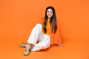 Fashionable and smiling brunette teenage girl in heels and denim jacket looking at camera while sitting and posing on orange background, cool and confident teenage girl, gen z fashion - obrazy, fototapety, plakaty