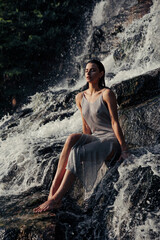 Young wet woman sitting on rock and enjoying with closed eyes near waterfall.