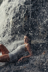 Young wet woman lying on rock near waterfall between water flows.