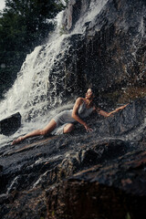 Young wet woman lying on rock near waterfall between water flows.