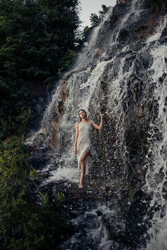 Young woman in wet dress standing near waterfall between water flows.