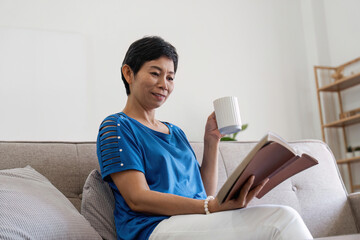 Serious confident aged female with cup of coffee sitting at desk with books literature at home