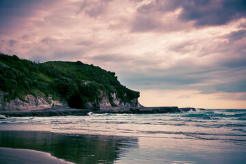 Sunset at Muriwai beach with waves splashing against the rocks and fishermen silhouettes in the distance. Auckland, New Zealand
