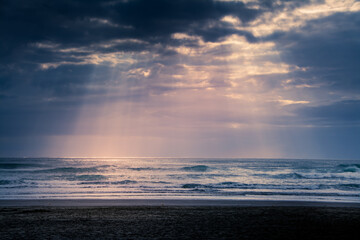 Sunset over Muriwai beach. Sun beams breaking through heavy clouds as waves splashing against the rocks. Auckland, New Zealand