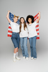 Fototapeta premium Full length of positive and multiethnic teenage girls in white t-shirts holding american flag and looking at camera on grey background, energetic teenage friends spending time, friendship