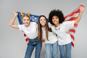 Joyful and multiethnic teenage girlfriends in casual white t-shirts and jeans looking at camera while holding american flag isolated on grey, energetic teenage friends spending time
