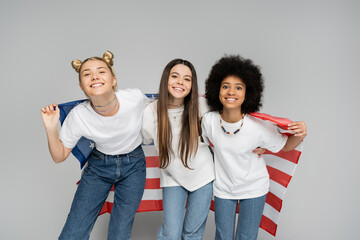 Joyful and interracial teenage girls in white t-shirts and jeans holding american flag and looking at camera while standing on grey background, lively teenage girls concept, friendship