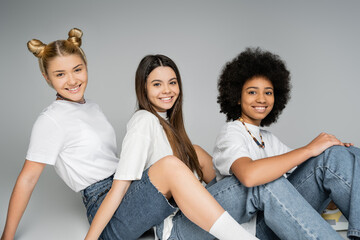 Joyful teen multiethnic girlfriends in white t-shirts and blue jeans looking at camera while sitting and posing together on grey background, multiethnic teen models concept, friendship and bonding