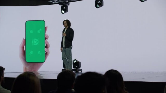 A man presenter is standing on a well-lit stage in front of a large LED screen showing a hand holding a generic smartphone with green screen. Technological, application or product launch presentation 