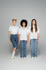 Naklejka premium Full length of lively and stylish multiethnic teenagers in white t-shirts and jeans looking at camera together while standing next to each other on grey background, trendy and stylish looks