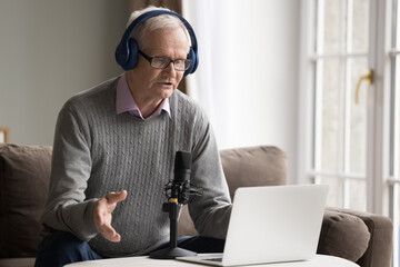Serious older man in headphones lead webinar or stream for internet audience subscribers, makes speech into microphone look at laptop screen, provide information, participate in virtual meeting event