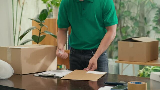 Mail worker in green uniform packing document in envelope and writing address on it before shipping to customer at office