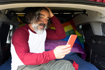 middle aged gay man sitting inside a camping van with lgtbi flag gesturing with a smartphone