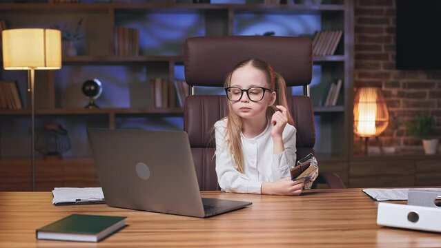 Precious little girl taking short break and eating delicious chocolate cookies in modern office. Pleasant kid in glasses keeping eyes closed and nodding head for expressing enjoyment.