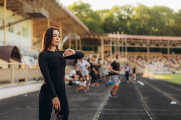 Fototapeta premium Female runner looks at smartwatch while going for run checks time listens music via headphones dressed in jacket strolls outdoor against modern tall buildings tracks wearable device wanders in city.