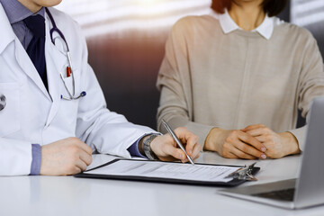 Fototapeta premium Unknown male doctor and patient woman discussing something while sittingin a darkened clinic and using clipboard, glare of light on the background