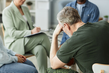 Focus on mature man puching his head while sitting in front of other people describing their problems to psychologist during session © Seventyfour