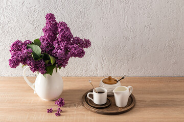 Spring still life. A jug with a bouquet of lilacs and a wooden round tray with a cup of morning coffee, a sugar bowl on a wooden countertop.