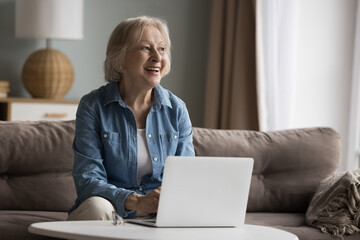 Attractive senior woman sits on sofa in living room looks thoughtful, rest with laptop at home,...