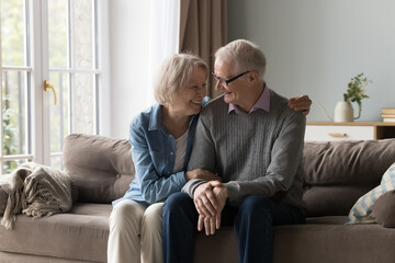 Cheerful attractive hoary mature spouses laughing, talking seated on couch in living room. Happy...