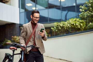 A smiling businessman pushing a bicycle and using a mobile phone, outdoors.