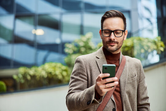A successful businessman with glasses on using a mobile phone outside the company.
