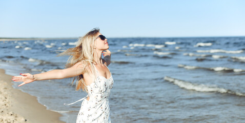 Fototapeta na wymiar Happy blonde beautiful woman on the ocean beach standing in a white summer dress and sun glasses, open arms