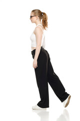 Fototapeta premium Side view of young blond woman in trousers and white blouse walking on white background