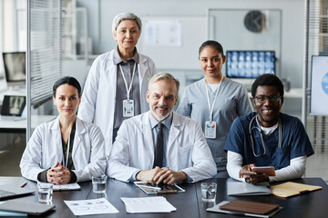 Group of successful professional clinicians in lab coats and uniform looking at camera with smiles while standing or sitting by workplace