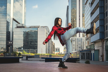 Photo shoot of stylish caucasian hip hop dancer posing kicking with red shirt, jeans and black boots shoes and top with a urban town as background. City shooting of moving model