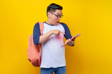 Surprised young Asian man student wearing t-shirt with backpack glasses pointing finger at book, exam on schedule isolated on yellow background. Education in high school university college concept