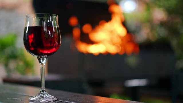 Close-up glass of red wine on the left with flame of burning fire burning at background reflecting in drink. Picnic on night backyard outdoors