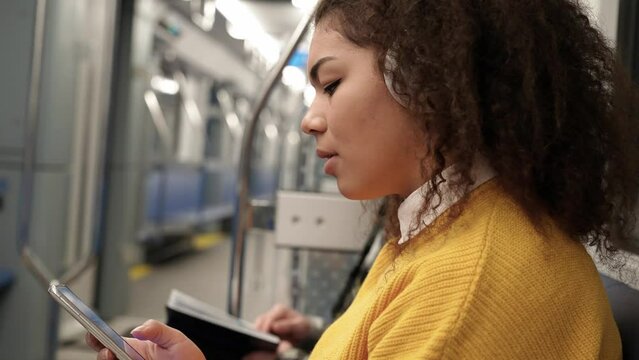 Young student ride the subway, dark skinned woman listen to music with headphones and uses a smartphones, surfs the internet, public transport.