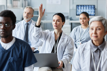 Young brunette general practitioner in lab coat raising hand after report of coach at seminar to...