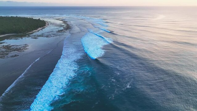 Drone shot tilting up from coral reef and island coastline with dense jungle and rolling waves, to the distant horizon at golden sunset