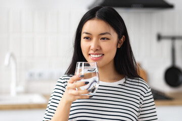 Beautiful Asian woman holding glass of clean fresh water standing in the home kitchen looks away...