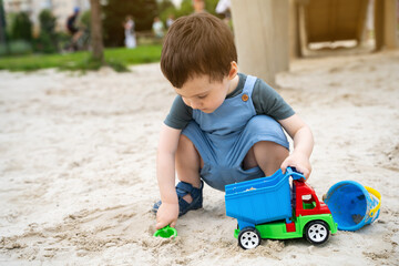 Little cute toddler boy 2.5 years old plays in the sandbox on a sunny summer day. Outdoor creative activities for kids. Toy car and shovel.