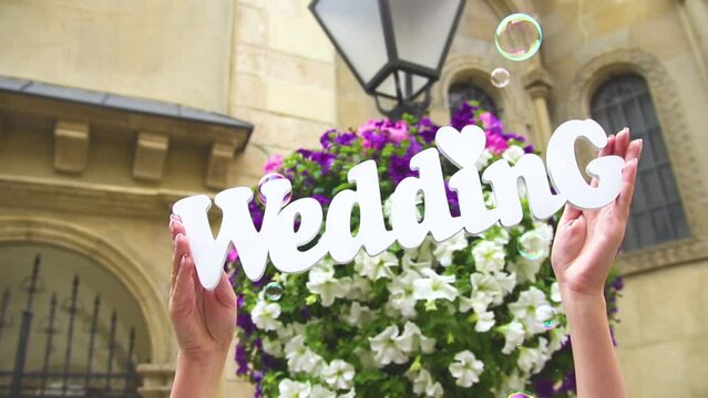 Young woman holding a white wooden word wedding in her hends above the head, on the background of a vase with colorful bright flowers. The wind blows the soap bubbles in the frame.