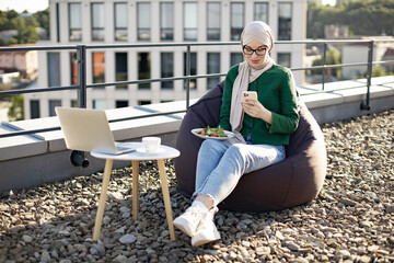 Young arabian woman in hijab holding phone while sitting near coffee table with laptop during lunch break on flat roof. Beautiful lady checking contact information before having bite of fresh salad.