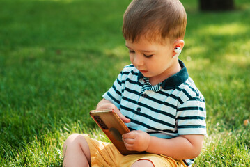 A cute European-looking boy studies the workings of a smartphone with wireless headphones in his ears on the green grass