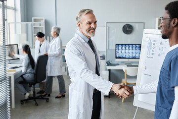 Mature experienced doctor congratulating young African American male colleague on successful speech at conference for medical personnel