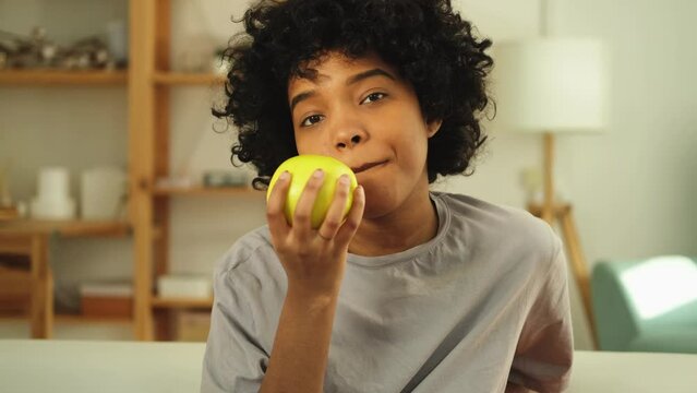 Happy pretty girl biting green apple at home. Beautiful african american young woman eating fresh fruit and smiling. Healthy food vegan vegetarian dieting concept. Healthy snack clean food