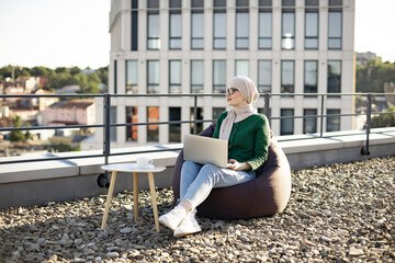 Beautiful woman wearing glasses and hijab daydreaming while holding laptop on knees in pouf chair outdoors. Arabian freelancer analyzing new project while staying on office roof terrace on sunny day.