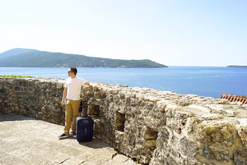 A young man, photographed full length, stands near a small suitcase near an old stone wall against the backdrop of the sea in a southern resort town.